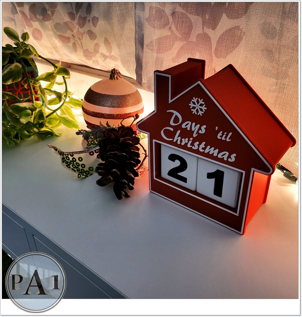 A red house-shaped calendar with the number 21 displayed, placed on a white surface next to a potted plant and a small decorative item.