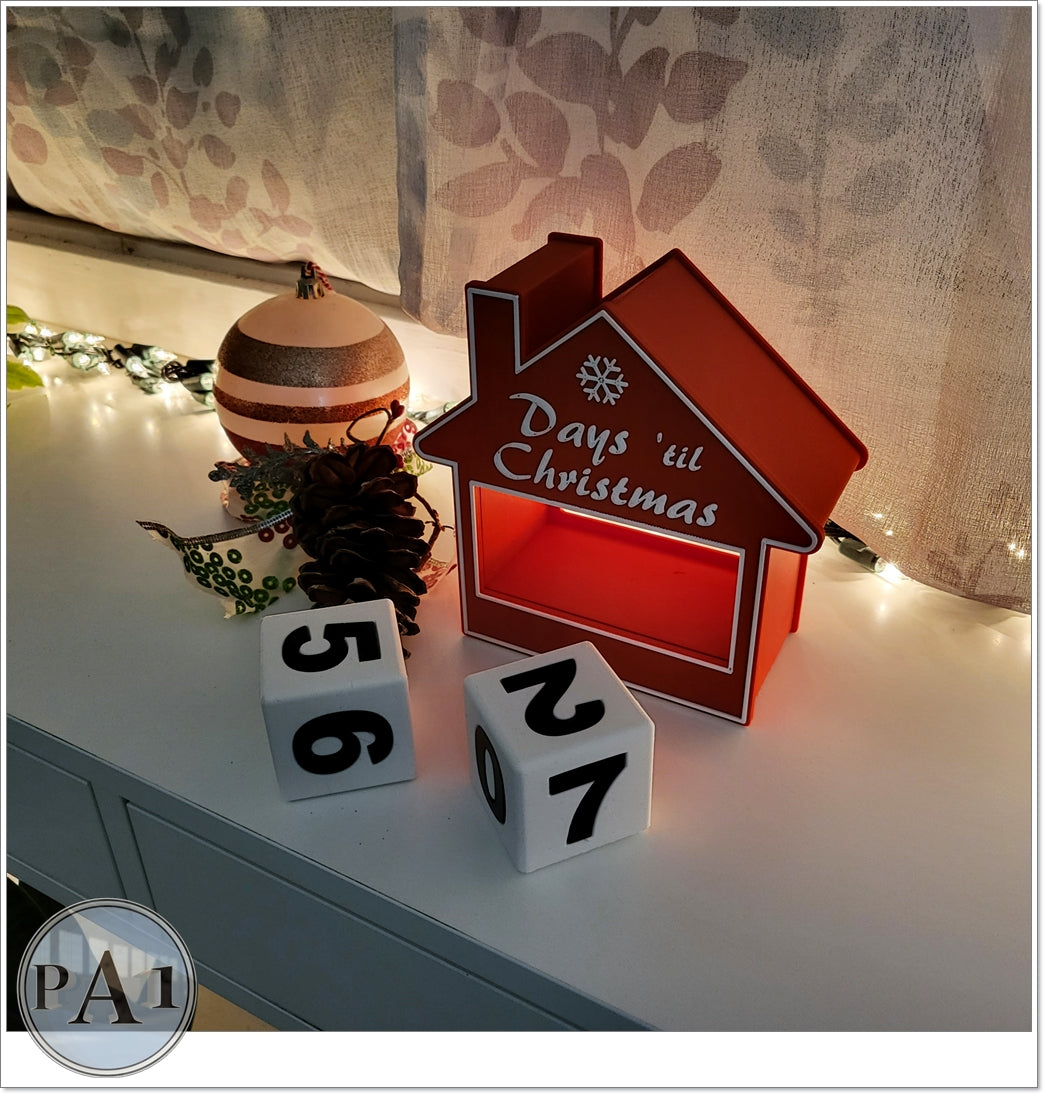 A red house-shaped box with the text "Days til Christmas" on it, along with two dice displaying the numbers "56" and "67" on a white surface.